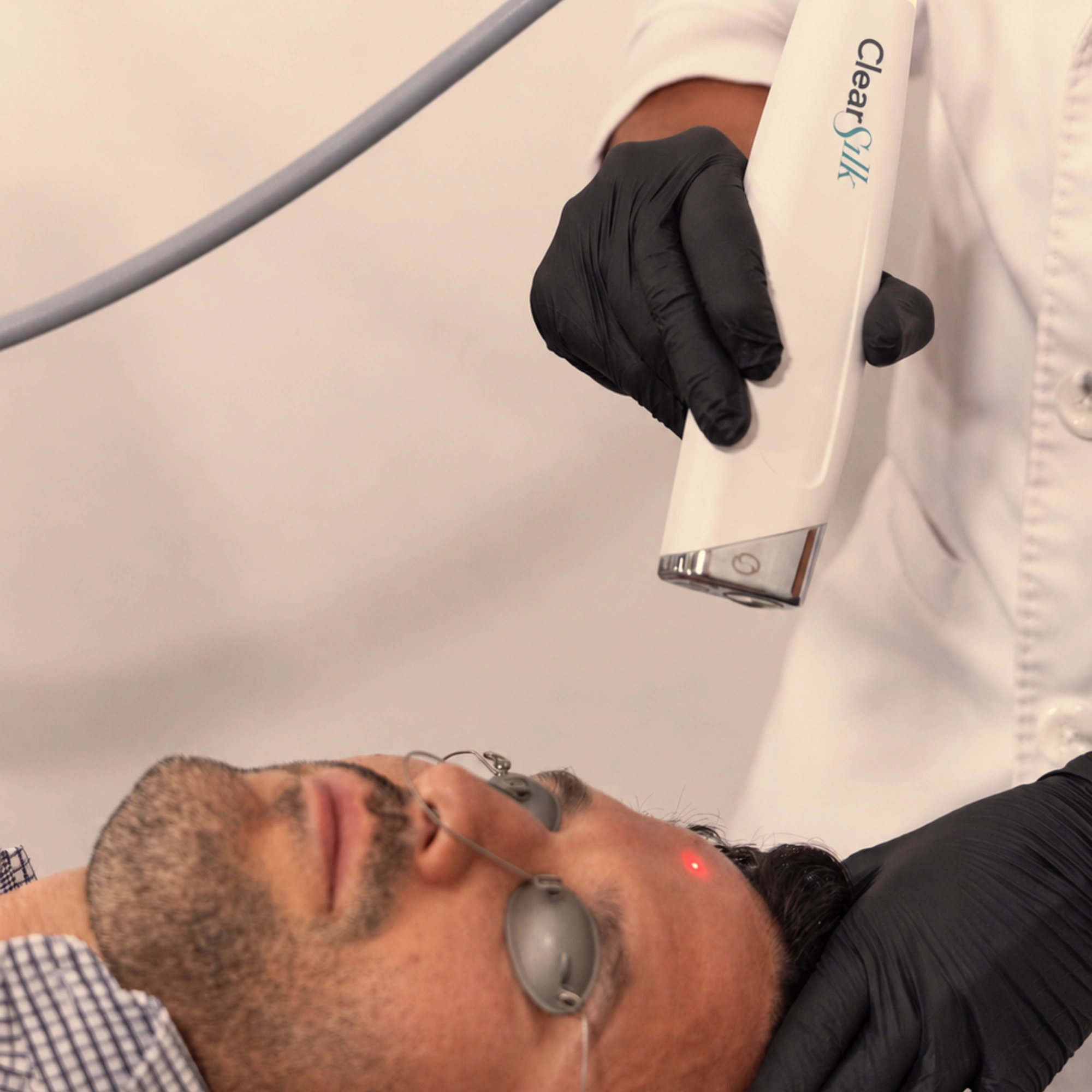 ClearSilk™ laser treatment performed on a man's forehead in a medical aesthetics clinic. The patient wears metal protective eye shields while the provider, wearing black gloves, applies the ClearSilk™ laser handpiece to gently rejuvenate the skin and improve overall skin quality.